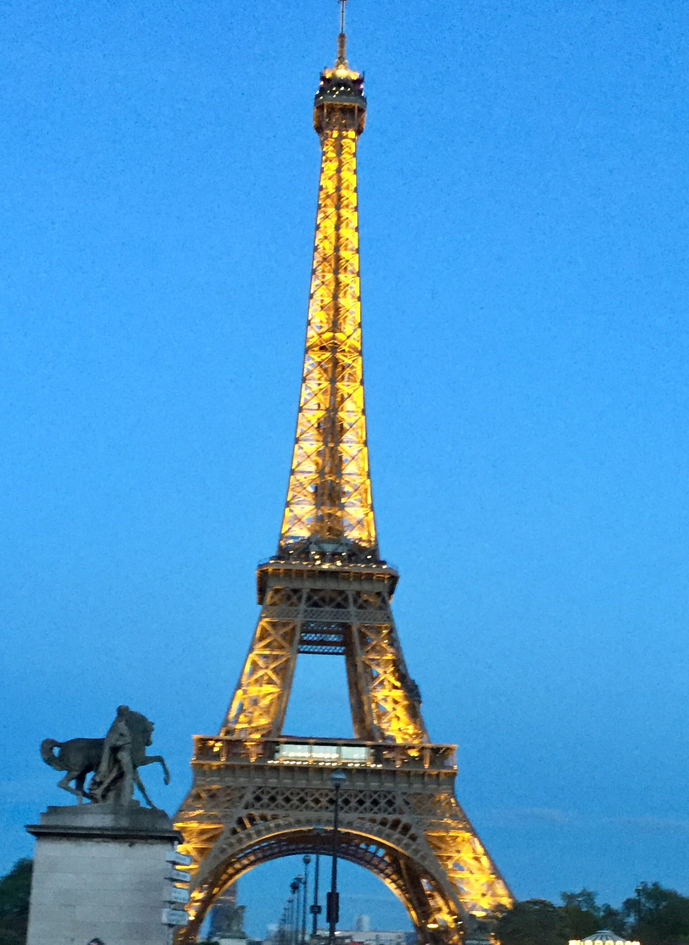 The Eiffel Tower glowing gold against a deep blue twilight sky, with a statue in the foreground